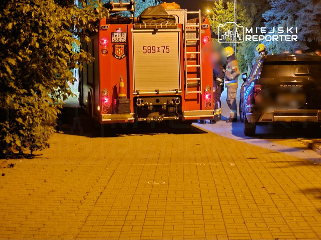 Strażacy w hełmach rozmawiają obok wozu strażackiego na oświetlonej ulicy, w tle widać zaparkowany samochód.