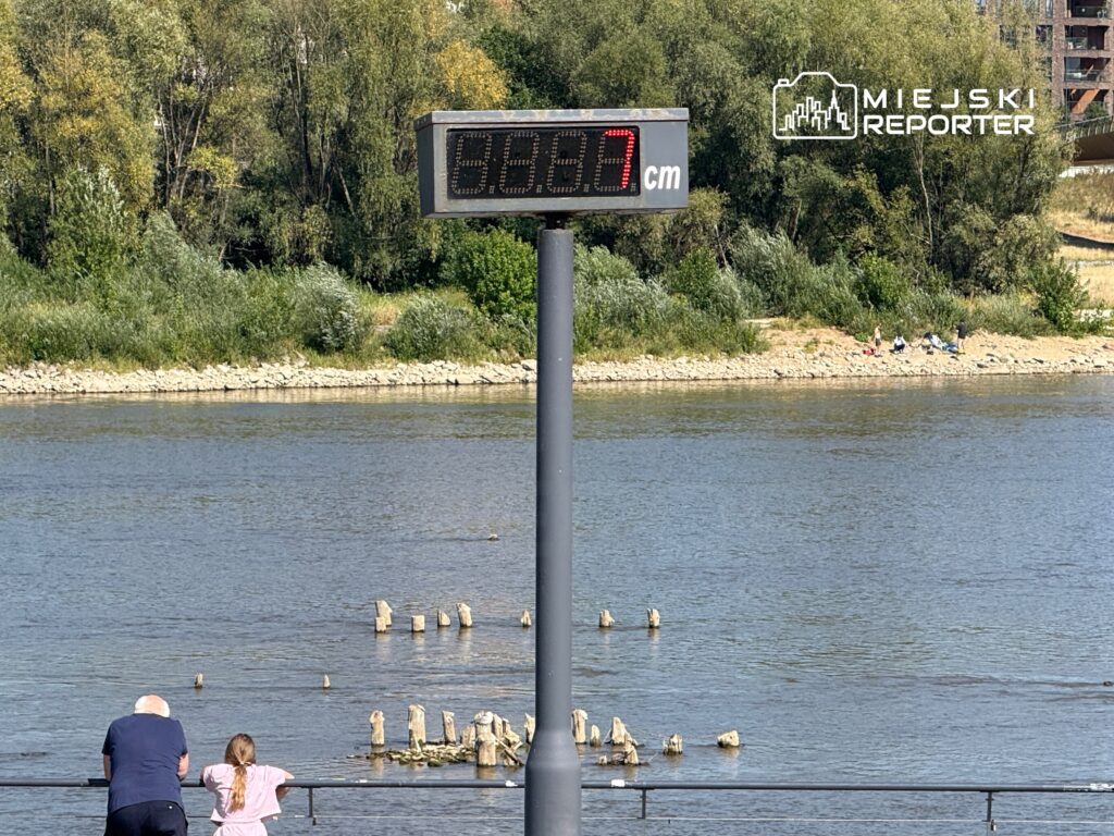 Dziecko w różowej koszulce i starszy mężczyzna obserwują rzekę, obok wyświetlacz z wysokością wody 68,7 cm.