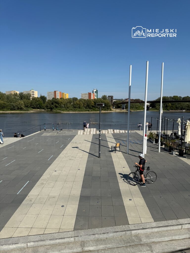 Mężczyzna w kasku trzymający rower na nowoczesnym deptaku nad rzeką, w tle kolorowe budynki.