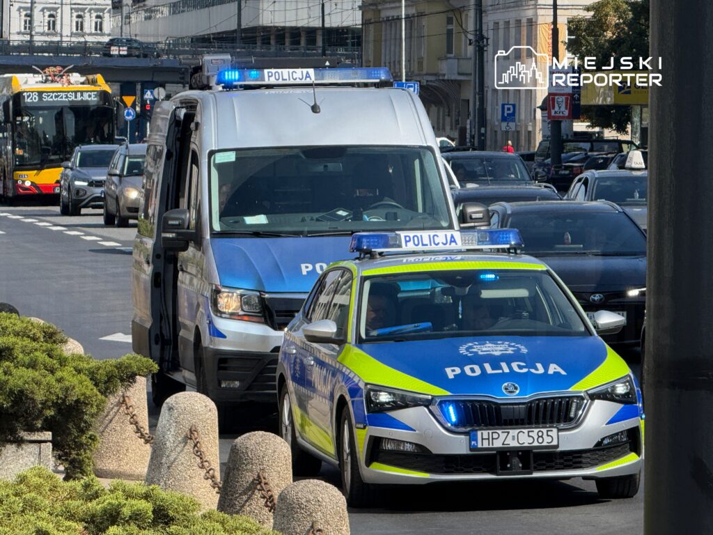 Radiowóz policyjny oraz van policyjny zatrzymane na ruchliwej ulicy, w tle autobus komunikacji miejskiej.