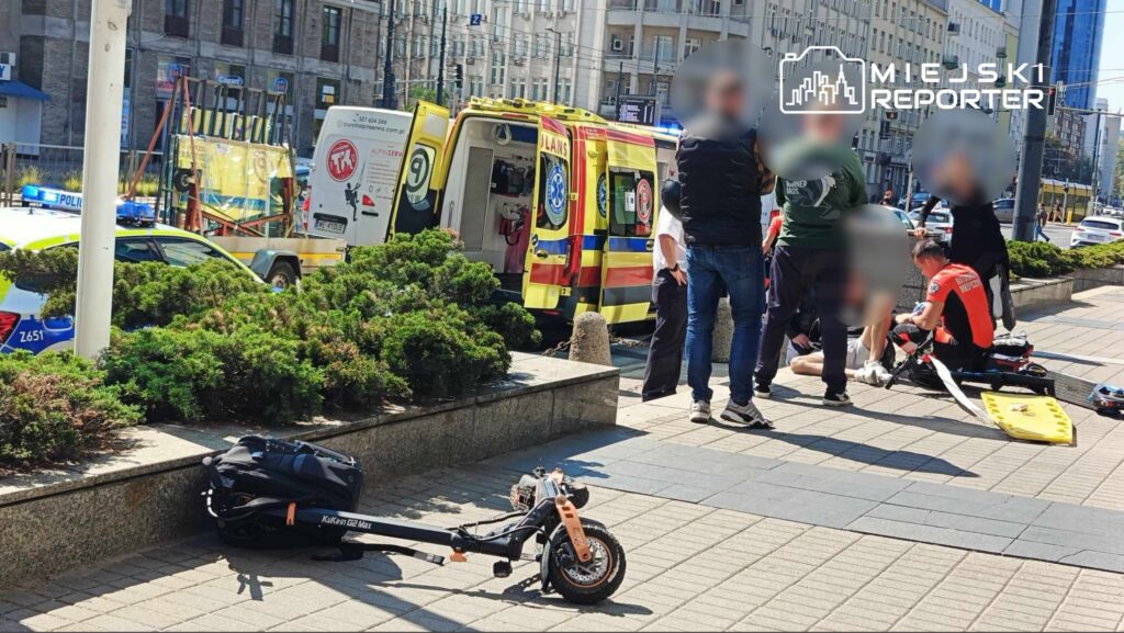 Grupa osób otacza rannego mężczyznę leżącego na chodniku, podczas gdy ratownicy medyczni udzielają mu pomocy. W tle widać ambulans oraz policyjny samochód.