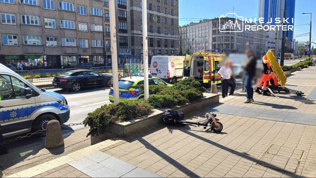 Na chodniku obok ulicy, zespół ratowników medycznych udziela pomocy poszkodowanemu, obok stoi ambulans.