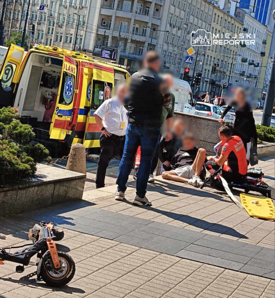 Grupa osób otacza mężczyznę leżącego na chodniku, podczas gdy ratownik medyczny udziela mu pomocy w pobliżu karetki.