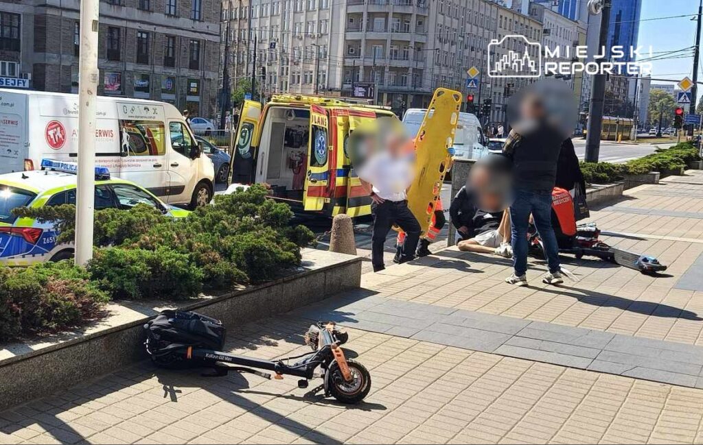 Grupa ratowników medycznych udziela pomocy osobie leżącej na chodniku obok przewróconego skutera.