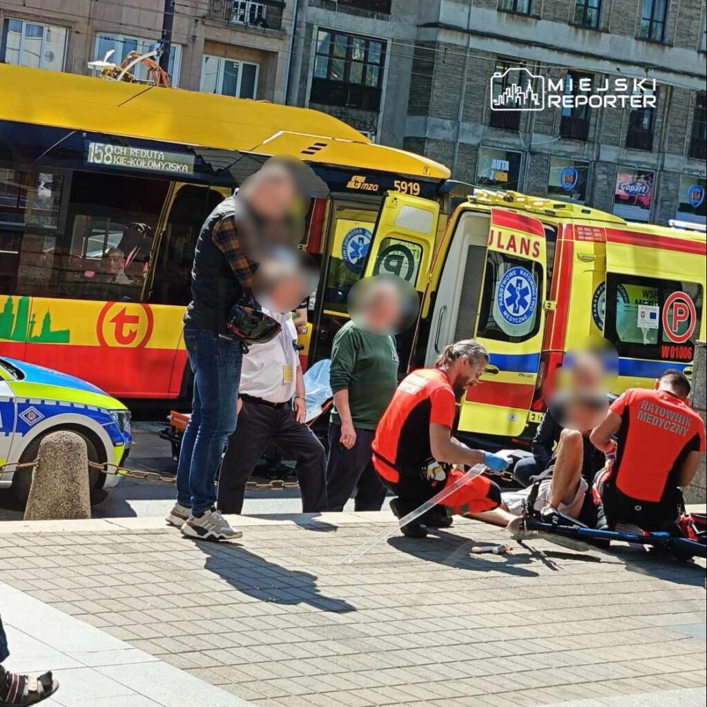 Ratownicy medyczni udzielają pomocy mężczyźnie na chodniku obok karetki i tramwaju w miejskim otoczeniu.