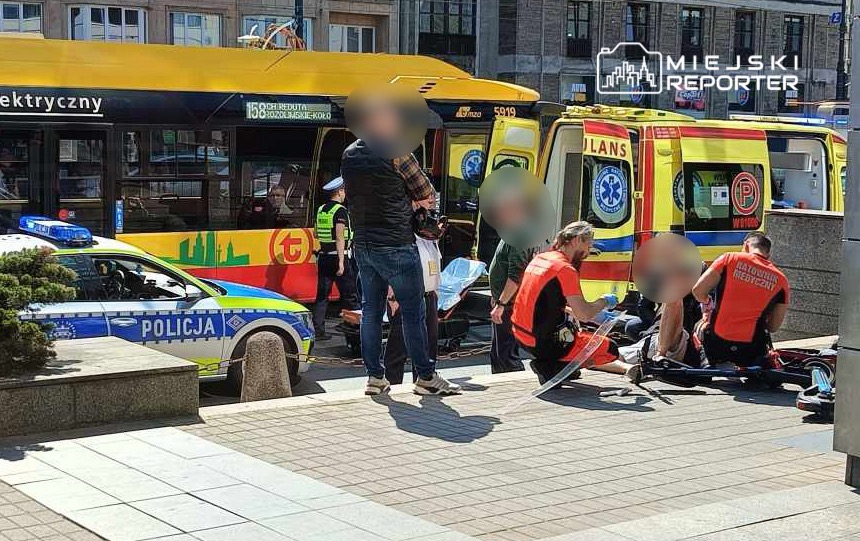 Ratownicy medyczni udzielają pomocy poszkodowanemu obok karetki i radiowozu na miejskiej ulicy.