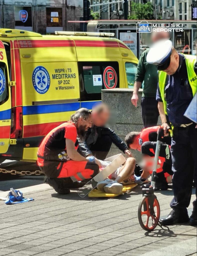 Ratownicy medyczni udzielają pomocy mężczyźnie leżącemu na chodniku, obok karetki pogotowia.