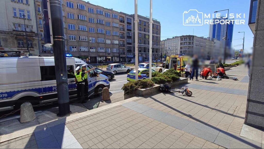 Policjanci w mundurach prowadzą interwencję obok radiowozu policyjnego, a w tle widać karetkę i grupę ratowników.