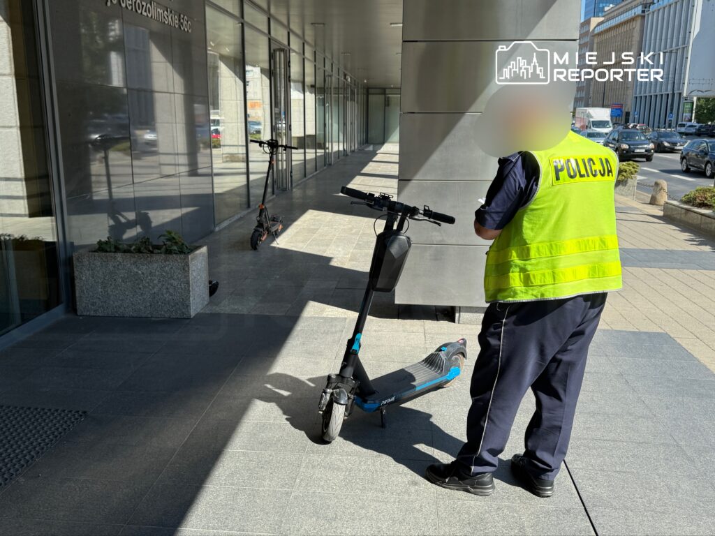 Policjant w kamizelce odblaskowej sprawdza elektryczną hulajnogę na chodniku przed nowoczesnym biurowcem.