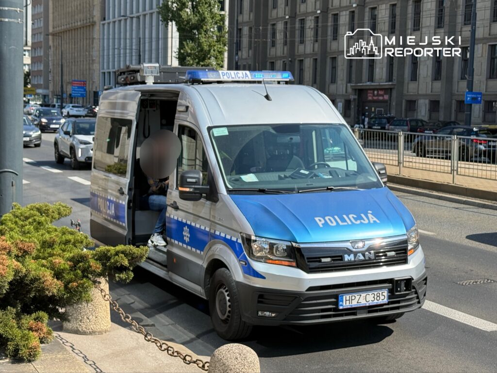 Policyjny radiowóz zaparkowany na ulicy, z otwartymi drzwiami, obok roślinności i przejeżdżającymi samochodami.