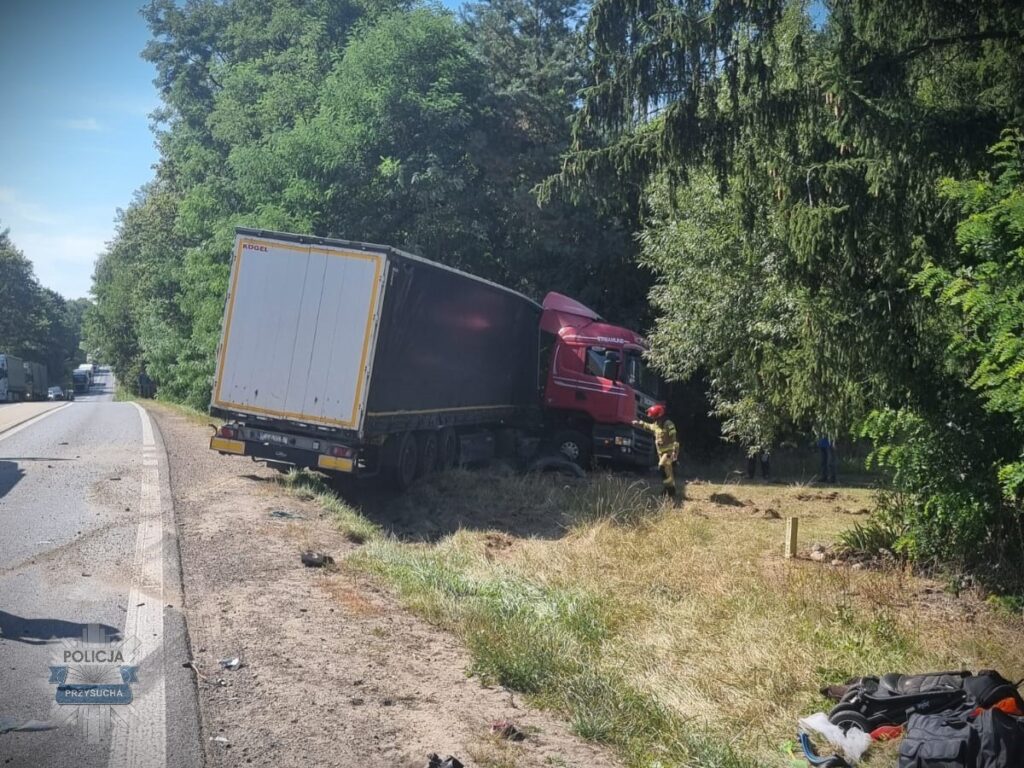 Czerwony ciężarówka w rowie przy drodze, obok strażak w hełmie przeszukujący teren. W tle widoczne inne pojazdy.