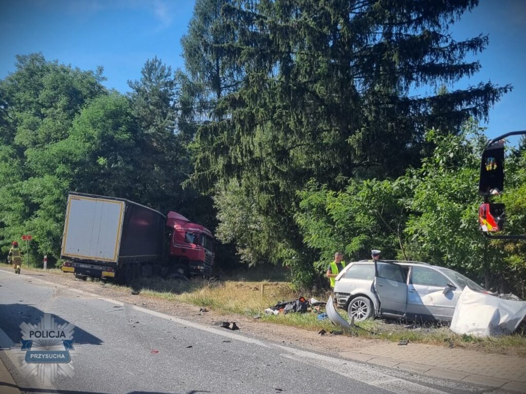 Czerwony ciężarowy samochód stoi na poboczu drogi, obok rozbite auto osobowe, a w tle pracują strażacy i policjanci.