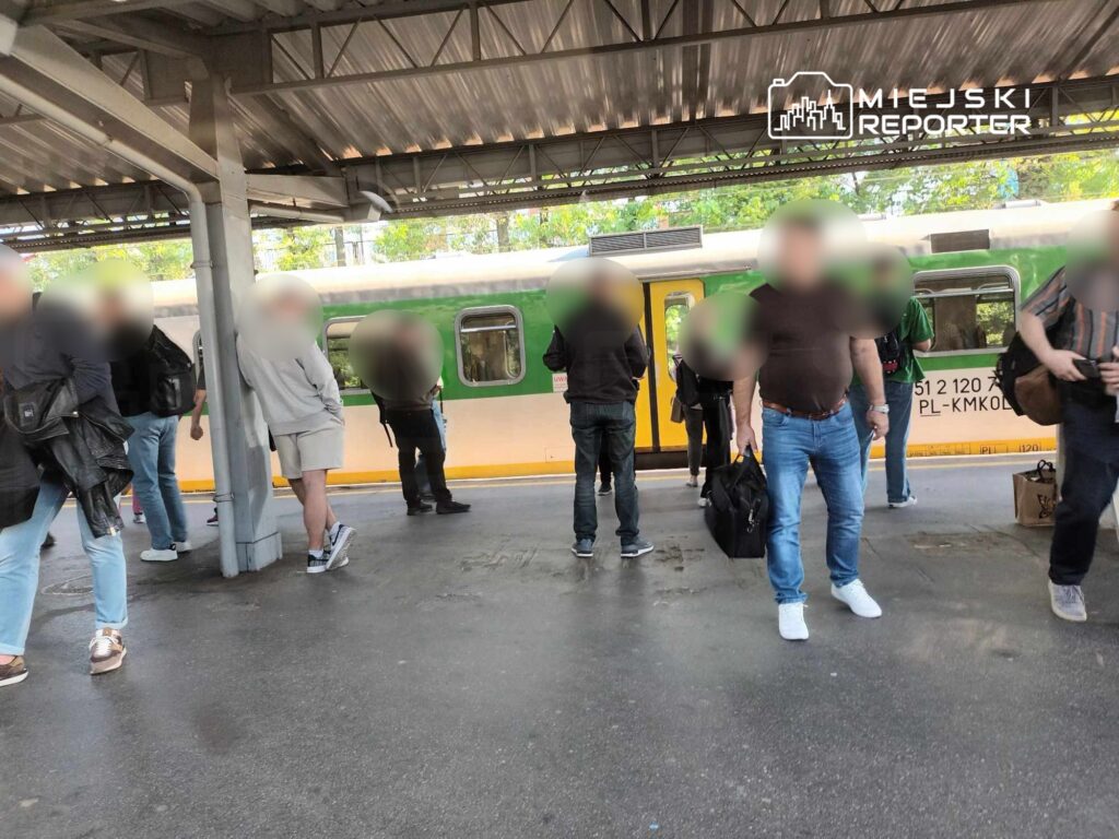 Grupa pasażerów czeka na peronie, obok zielono-żółtego pociągu, w otoczeniu zadaszonej stacji.