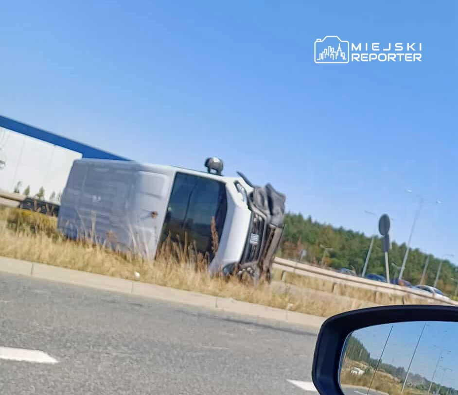 Biały samochód dostawczy leży na boku na poboczu drogi, w tle widoczne są inne pojazdy i zieleń.