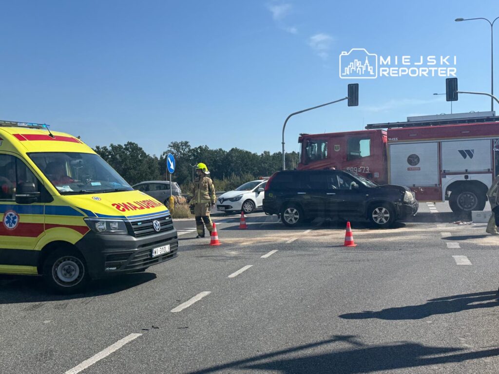 Ambulans i wóz strażacki na miejscu wypadku, strażak kierujący ruchem w pobliżu uszkodzonego pojazdu.