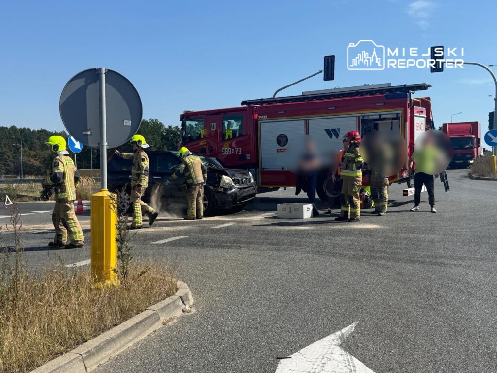 Czterech strażaków w kaskach i mundurach zabezpiecza miejsce wypadku drogowego, obok uszkodzonego samochodu.
