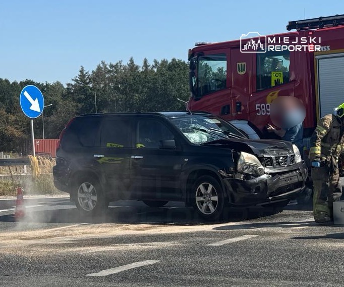 Czarny samochód osobowy z uszkodzoną przednią częścią stoi na drodze, obok niego strażak w mundurze.