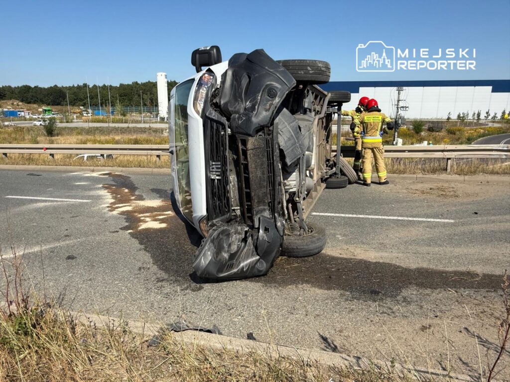 Przewrócony samochód osobowy leży na boku na poboczu drogi, obok interweniują strażacy w mundurach.