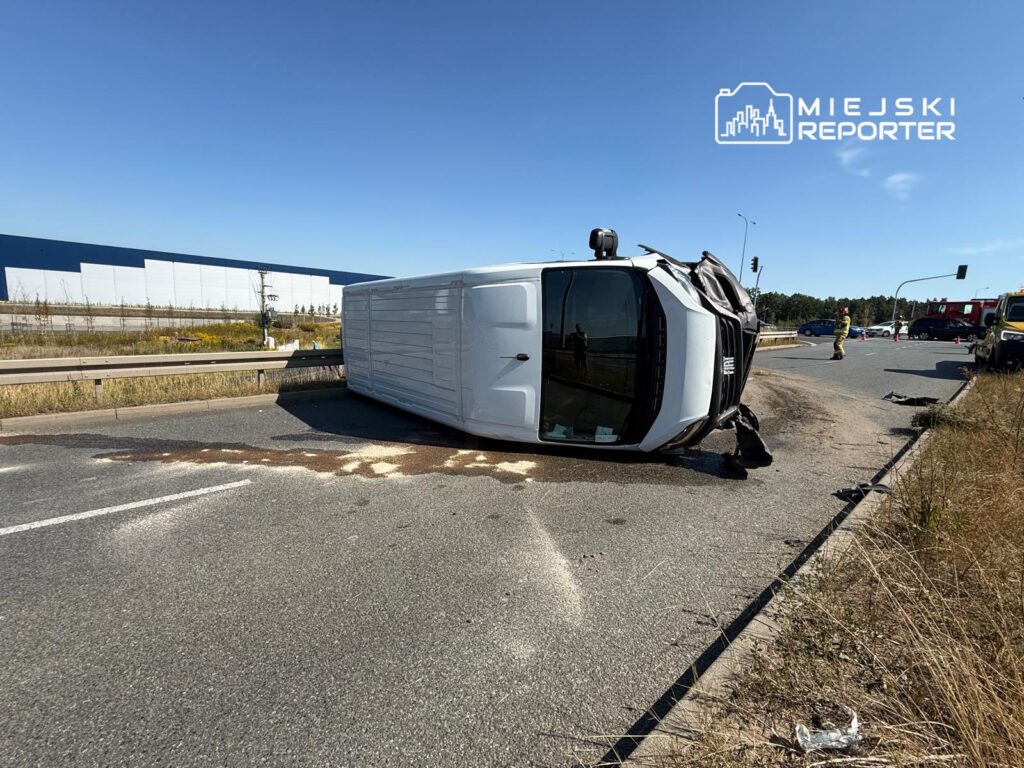 Przewrócony dostawczy samochód leży na drodze, obok interweniują strażacy, w tle nowoczesny budynek.