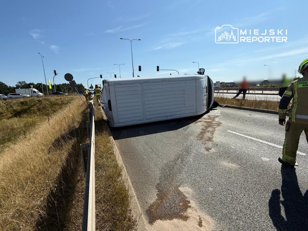 Na drodze leży przewrócony samochód dostawczy, a obok pracuje dwóch ratowników. W tle ruch uliczny.
