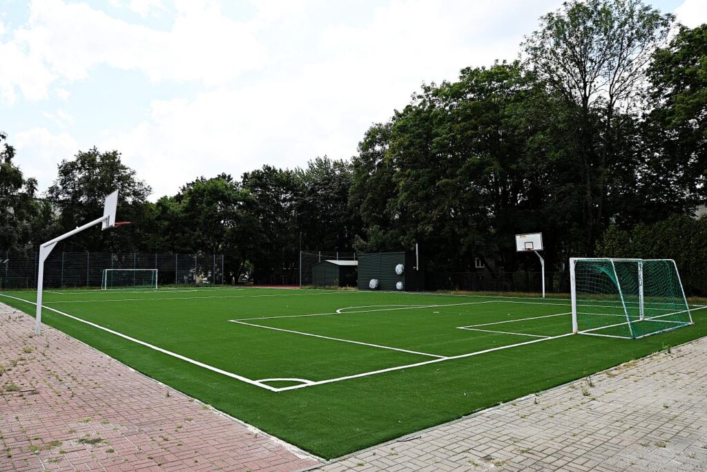 Nowoczesne boisko sportowe z sztuczną trawą, bramkami piłkarskimi i koszami do koszykówki, otoczone drzewami.