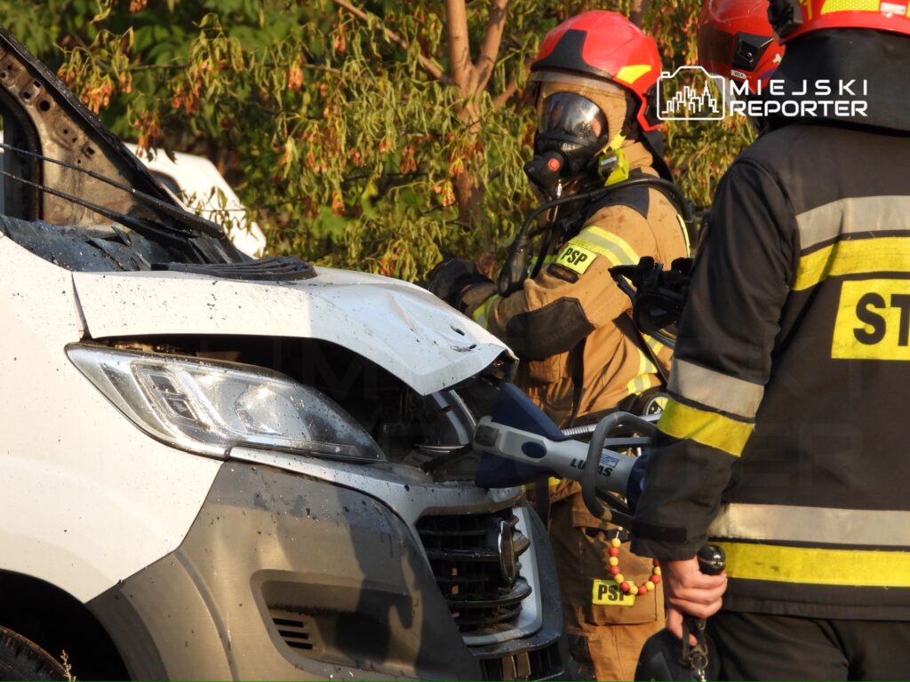 Strażacy w sprzęcie ochronnym pracują nad rozbitym pojazdem, usuwając uszkodzenia w otoczeniu zieleni.