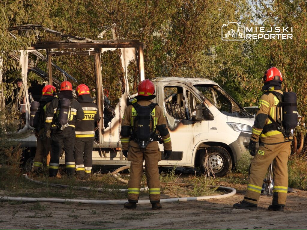 Grupa strażaków w hełmach obserwuje spaloną furgonetkę w zaroślach, z wężem strażackim na ziemi.