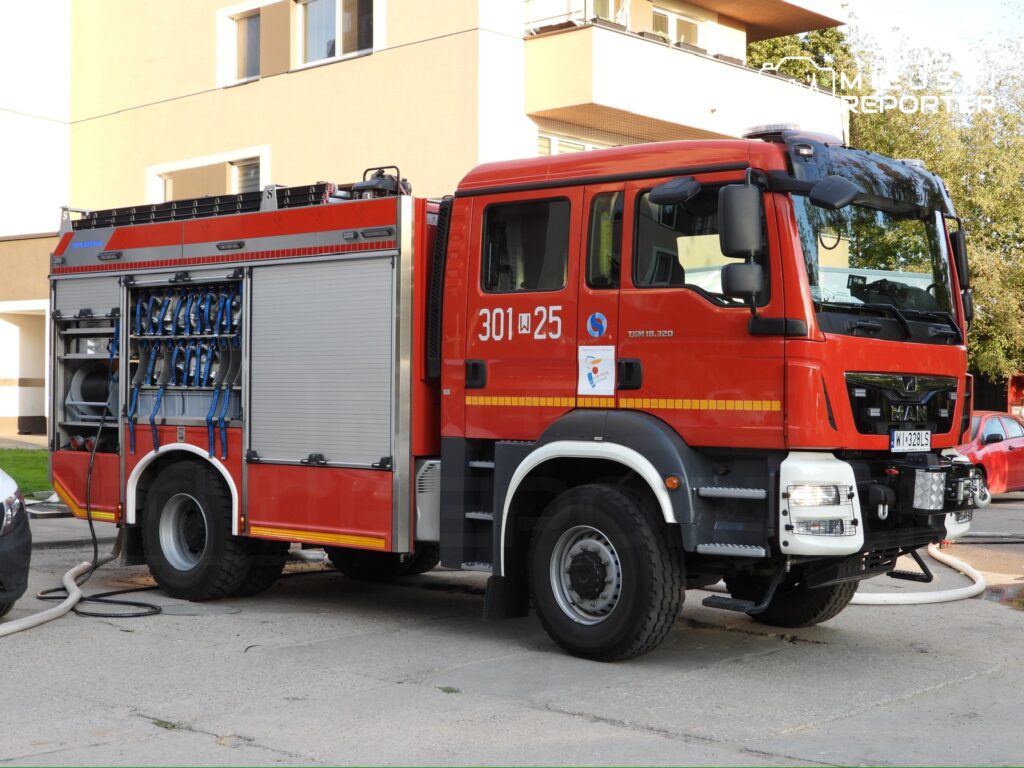 Czerwony wóz strażacki zaparkowany na chodniku obok budynku mieszkalnego, z wężami wodnymi podłączonymi do hydrantu.