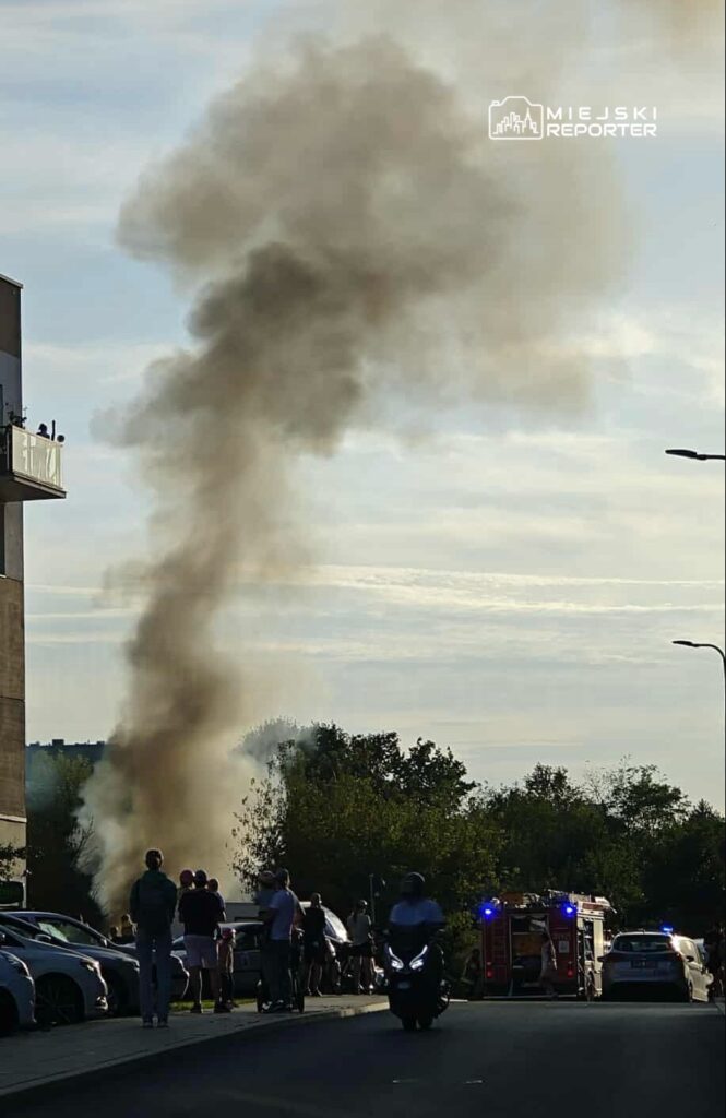 Grupa ludzi obserwuje dym unoszący się z miejsca pożaru w miejskim otoczeniu, w pobliżu samochodów i wozów strażackich.