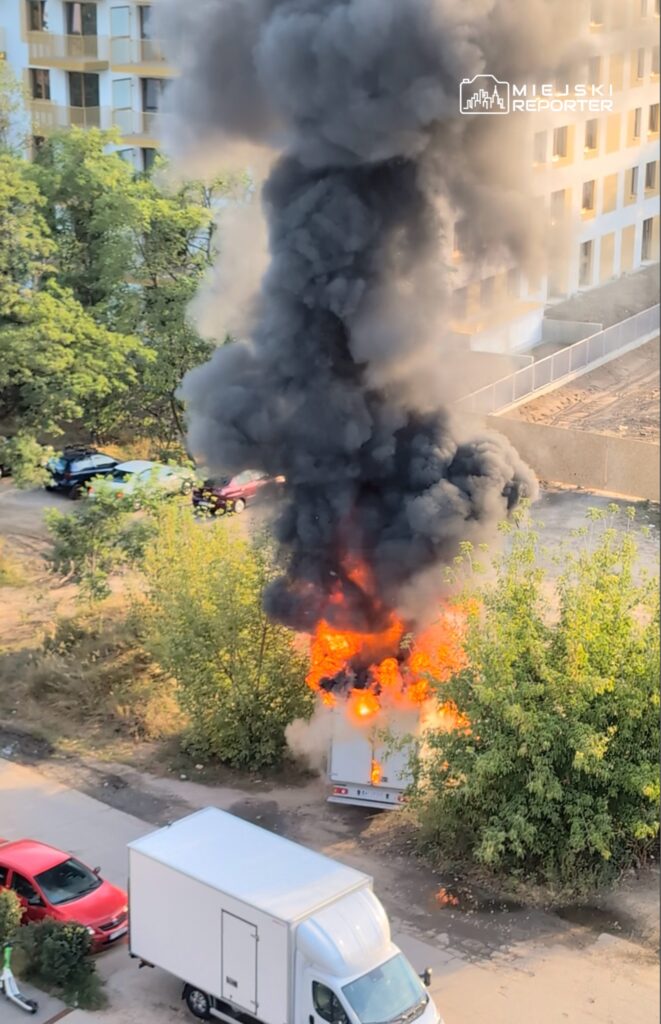 Pojazd dostawczy płonący na parkingu, wydobywający gęsty, czarny dym i płomienie w otoczeniu drzew.