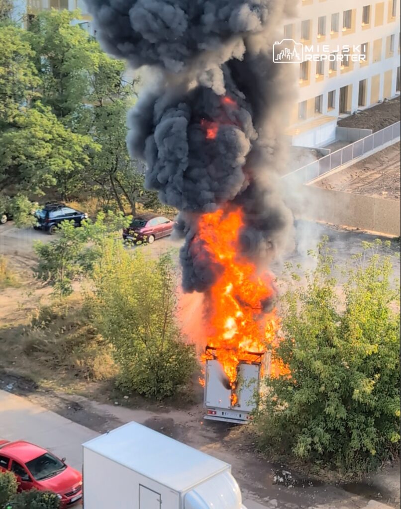 Pojazd dostawczy w płomieniach, unoszący się czarny dym w otoczeniu drzew i zaparkowanych samochodów.