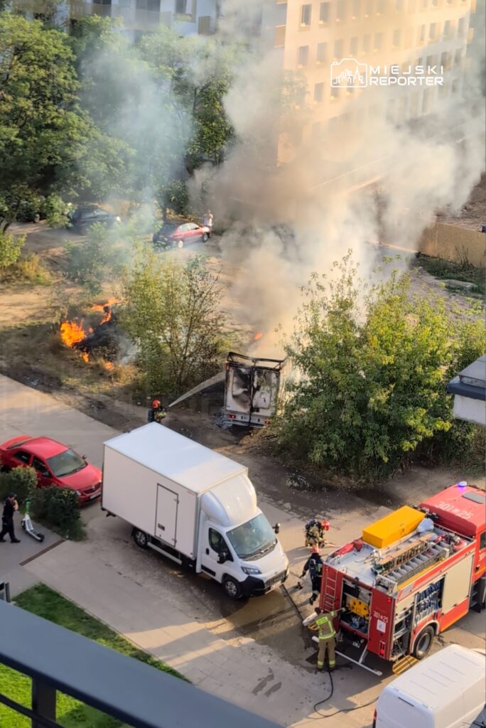 Strażacy gaszą pożar w pobliskim busie, obok widoczne czerwone samochody oraz dym unoszący się z terenu.