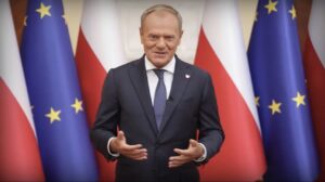 Mężczyzna w garniturze uśmiecha się i gestykuluje przed flagami Polski oraz Unii Europejskiej.