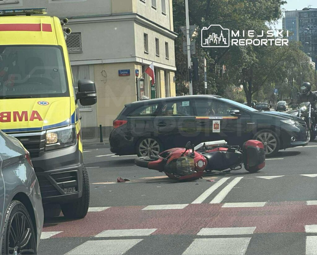 Ambulans zatrzymany przy przewróconym skuterze na ulicy, obok przejeżdżające samochody.