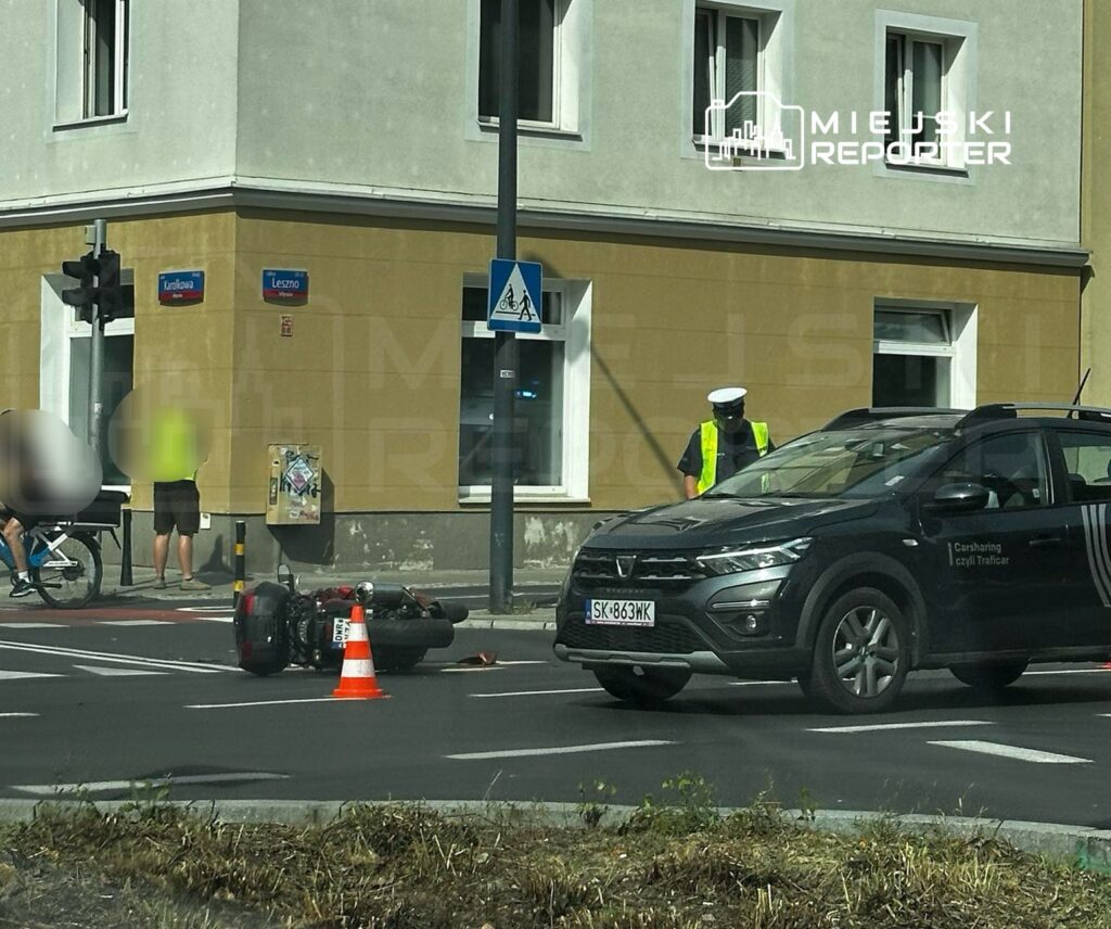 Motocykl leży na jezdni obok pomarańczowego pachołka, a policjant w kamizelce odblaskowej rozmawia z przechodniami.