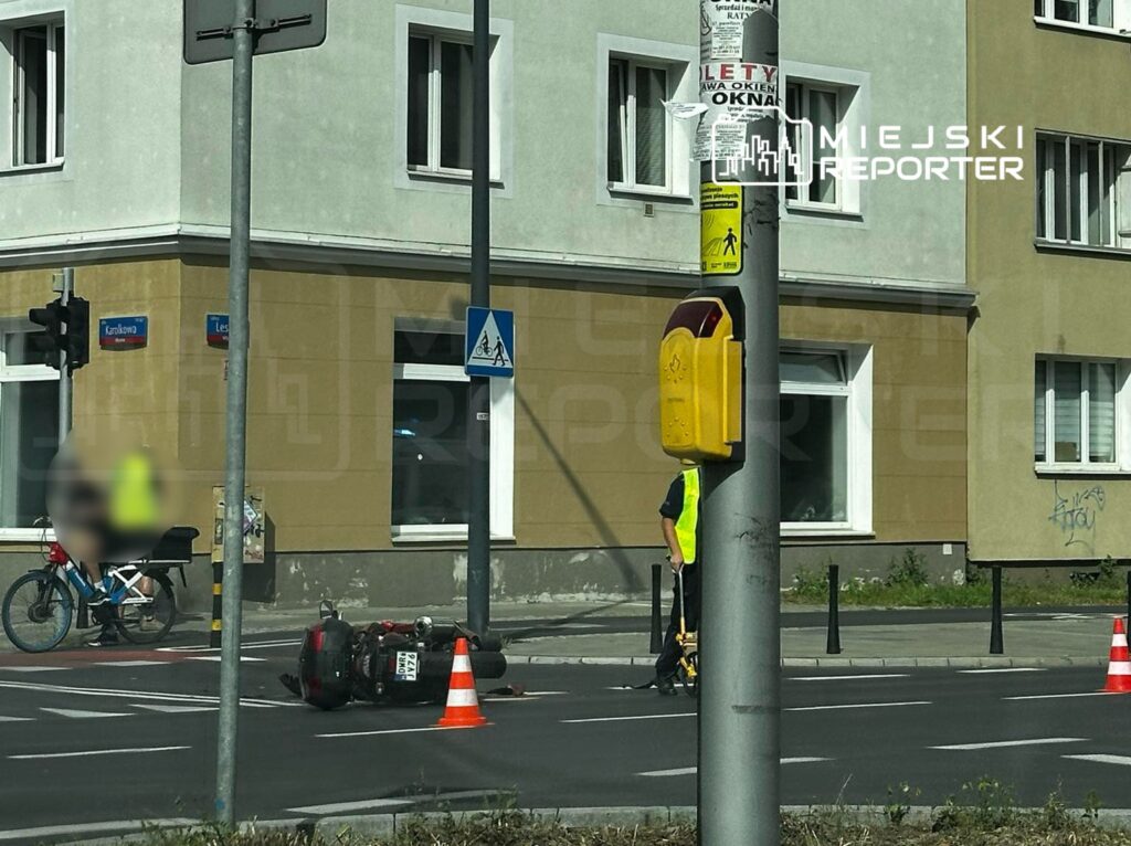 Motocykl leży na boku na jezdni, a obok stoi policjant w kamizelce odblaskowej, kierujący ruchem.
