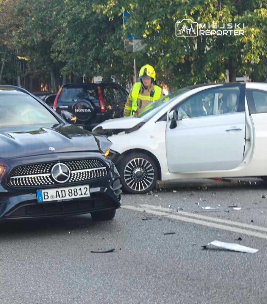 Mężczyzna w hełmie ochronnym bada uszkodzone samochody po kolizji na ulicy, obok leżą fragmenty wozów.