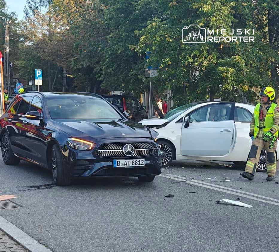 Czarny samochód osobowy z przodu, obok uszkodzony biały pojazd, a obok nich strażak w mundurze, zbierający szczątki.