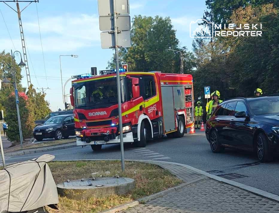 Czerwony wóz strażacki z włączonymi sygnałami świetlnymi stoi na skrzyżowaniu, obok grupy strażaków w odblaskowych kamizelkach.