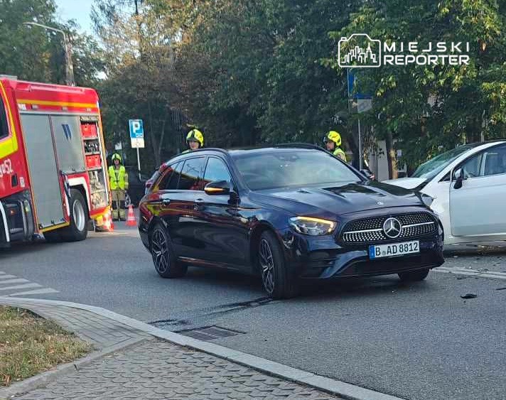 Czarny samochód osobowy zaparkowany obok uszkodzonego białego auta, w tle strażacy w kaskach przy wozie strażackim.