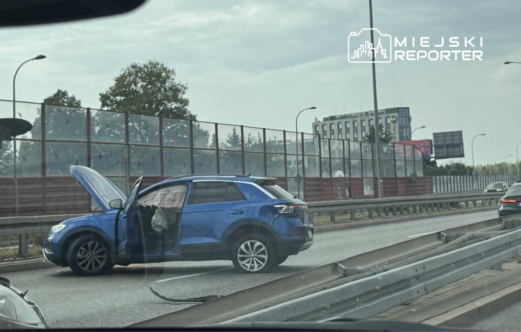 Niebieski SUV zatrzymany na pasie ruchu, z otwartą maską i drzwiami, obok ogrodzenie drogi.