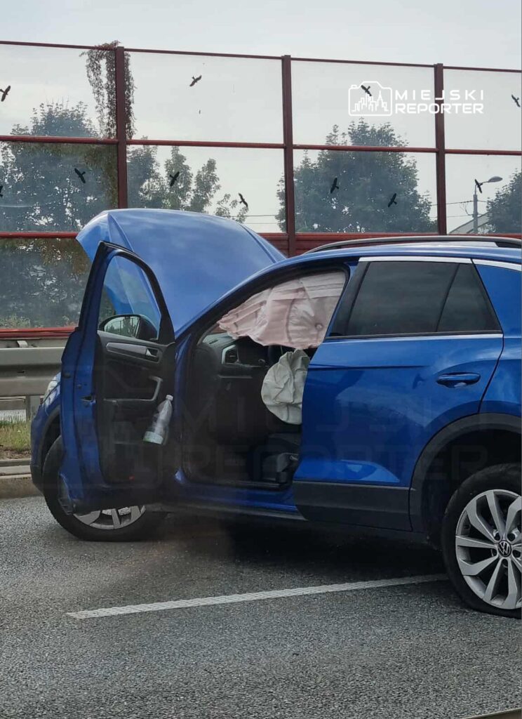 Niebieski samochód osobowy z otwartymi drzwiami i uniesioną maską stoi na poboczu drogi. Wewnątrz widoczny jest koc oraz butelka.
