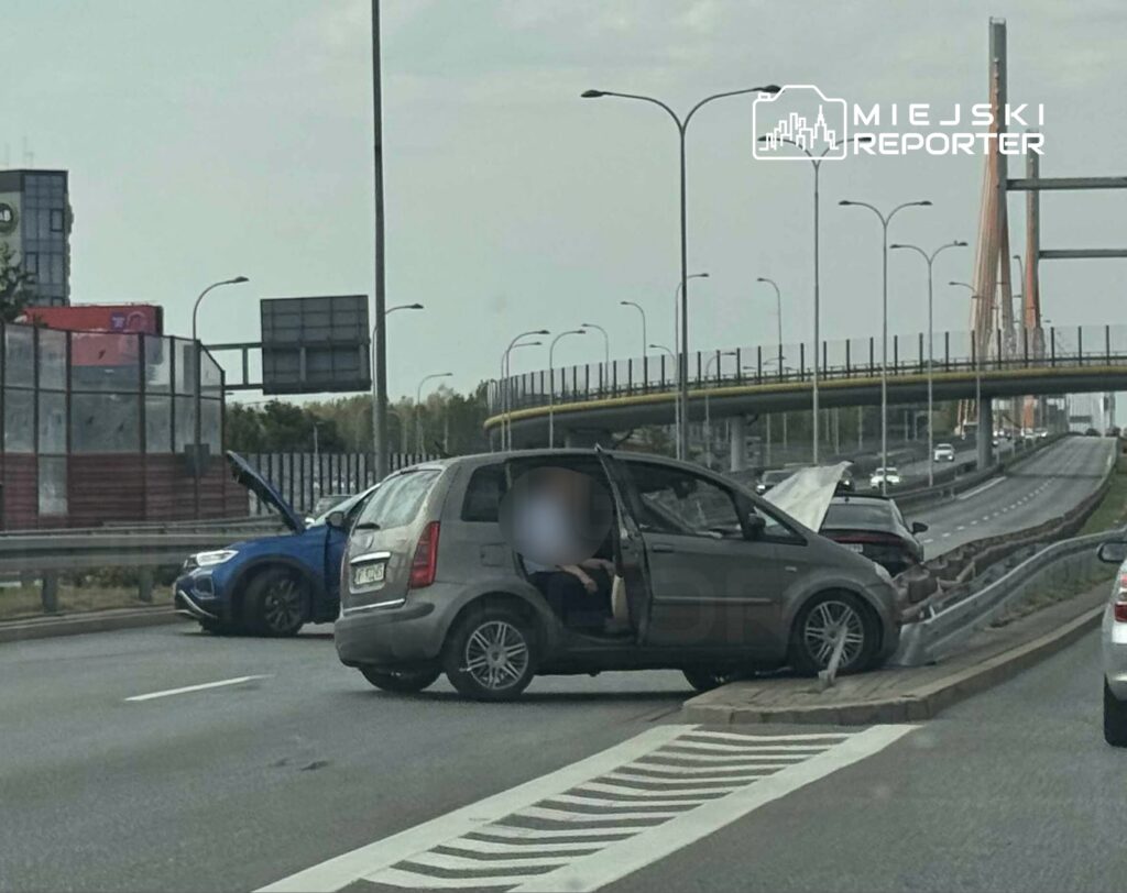 Dwa uszkodzone samochody na pasie drogowym, jeden z otwartą maską, w tle widoczny most.