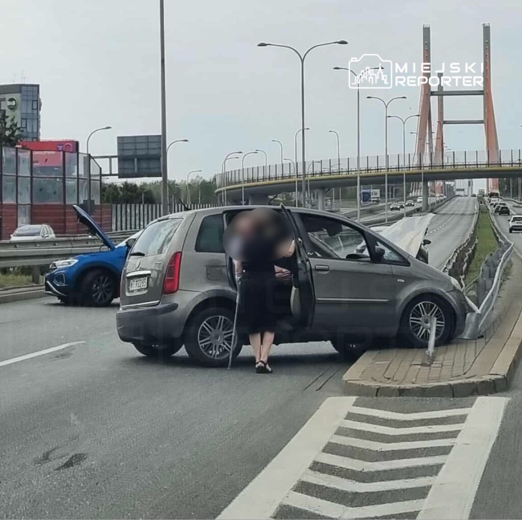 Kobieta z laską stoi obok otwartego samochodu na pasie ruchu, a w tle widać most i inne pojazdy.