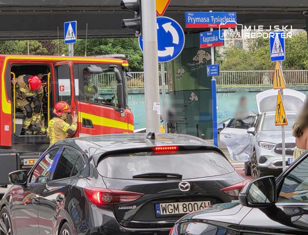 Strażacy w specjalnych strojach wchodzą do wozu strażackiego na ruchliwej ulicy, obok uszkodzonego samochodu.