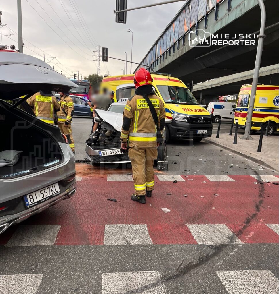 Strażacy badają uszkodzony samochód osobowy na skrzyżowaniu, obok stoją karetki pogotowia.