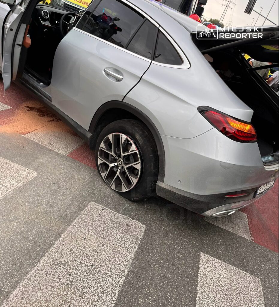 Srebrny SUV z przebitą oponą stoi na przejściu dla pieszych, obok otwartych drzwi kierowcy.