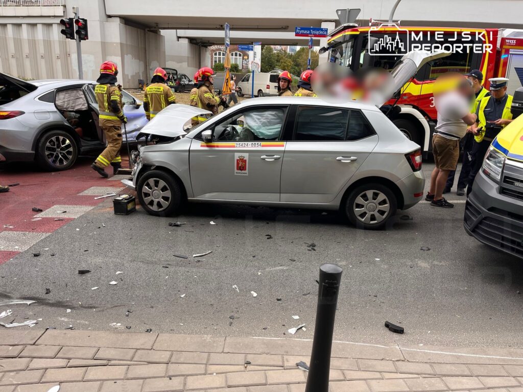 Na skrzyżowaniu ulic leżą uszkodzone samochody, a strażacy i policjanci udzielają pomocy poszkodowanym.