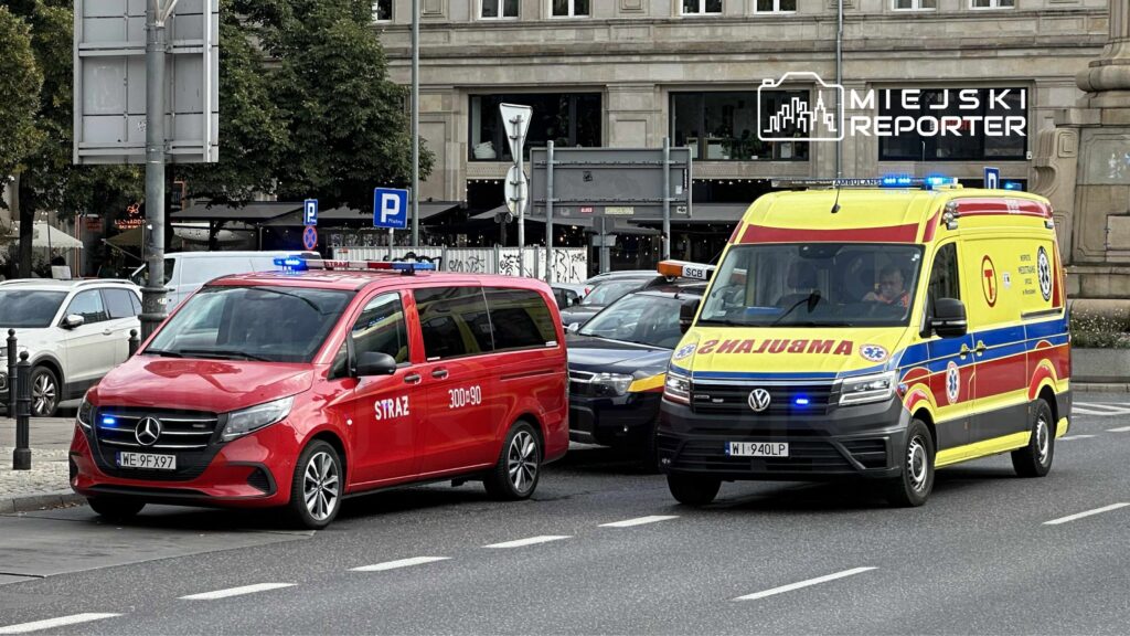 Czerwony samochód strażacki i żółta karetka przejeżdżają przez ruchliwą ulicę w mieście, z włączonymi sygnałami świetlnymi.