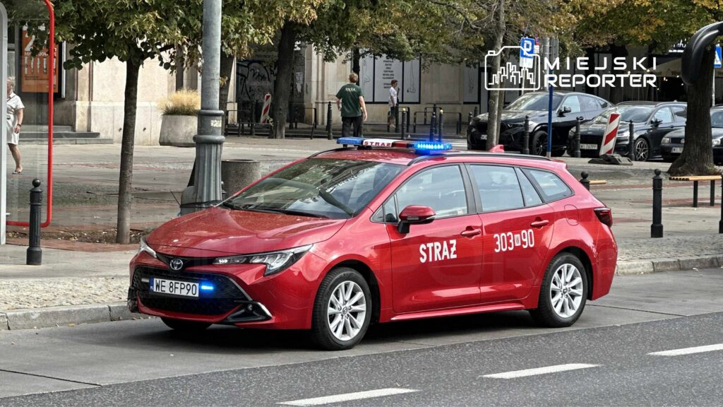 Czerwony samochód straży miejskiej z włączonymi sygnalizatorami świetlnymi parkuje na ulicy w miejskim otoczeniu.